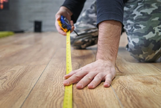 Homme en tenue de camouflage agenouillé sur parquet en bois, utilisant un mètre ruban jaune pour mesurer une surface au sol dans un intérieur résidentiel